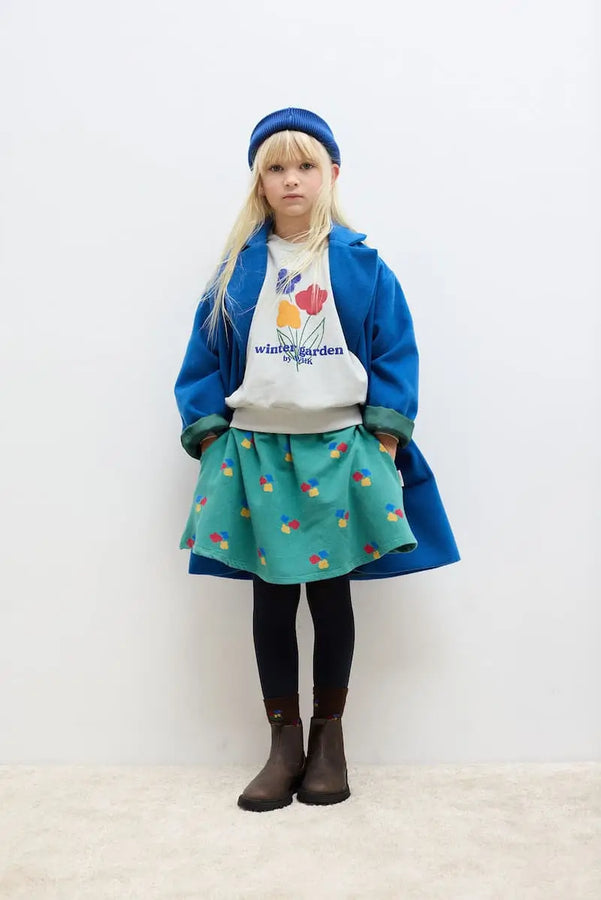 Niña con falda verde con estampado de flores, sudadera gris y abrigo largo azul, todo de Weekend House Kids moda sostenible.