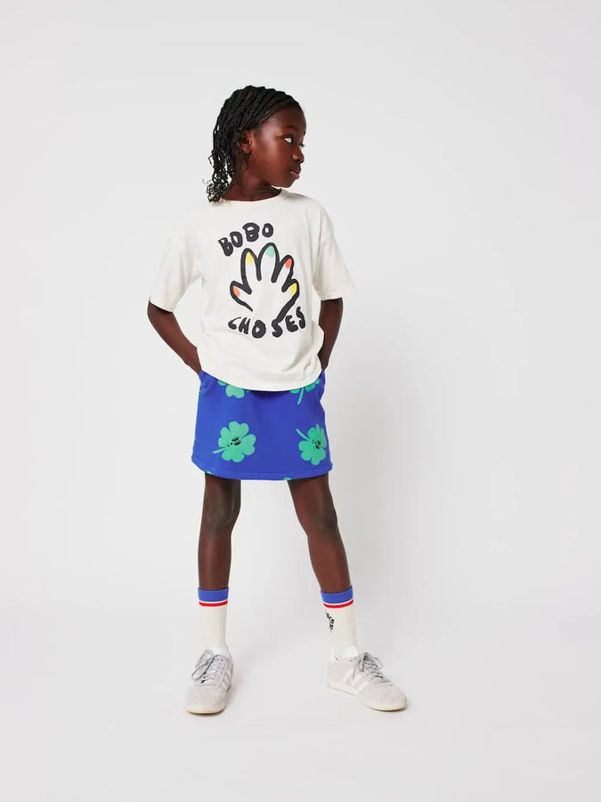 Niña lleva una falda corta azul de Bobo Choses con estampado de tréboles verdes conjuntada con una camiseta de color blanco, calcetines blancos y deportivas grises. 