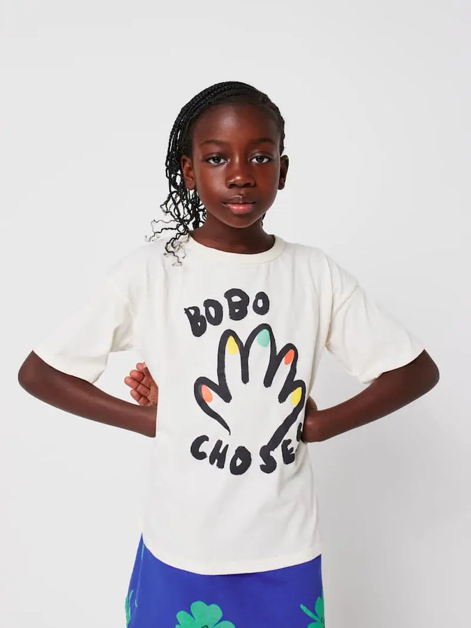 Niña vistiendo camiseta blanca con dibujo de mano con uñas multicolores y falda azul con tréboles, ambas de Bobo Choses.