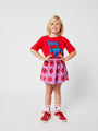 Niña de frente con falda corta de Bobo Choses color morado con estampado de caleidoscopios rojos, con camiseta y zapatillas rojas a juego. 