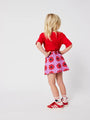 Niña de espaldas con falda corta de color morado de Bobo Choses con estampado de caleidoscopios rojos, con camiseta y zapatillas rojas a juego. 