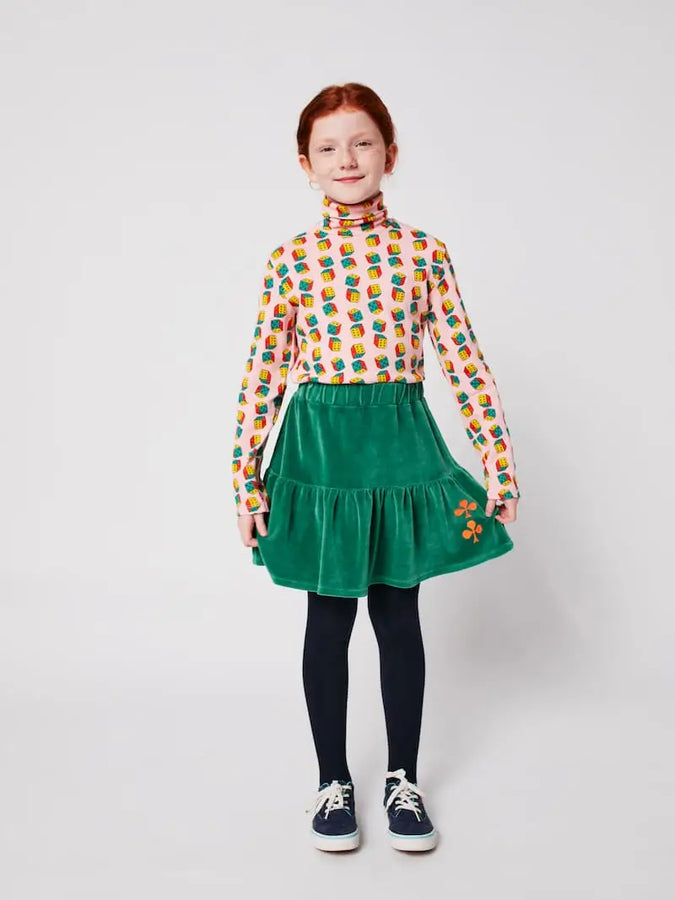 Niña con falda corta verde de terciopelo para niñas de Bobo Choses con volante y tréboles bordados en color tierra, y con top de cuello alto con estampado de dados multicolores. 