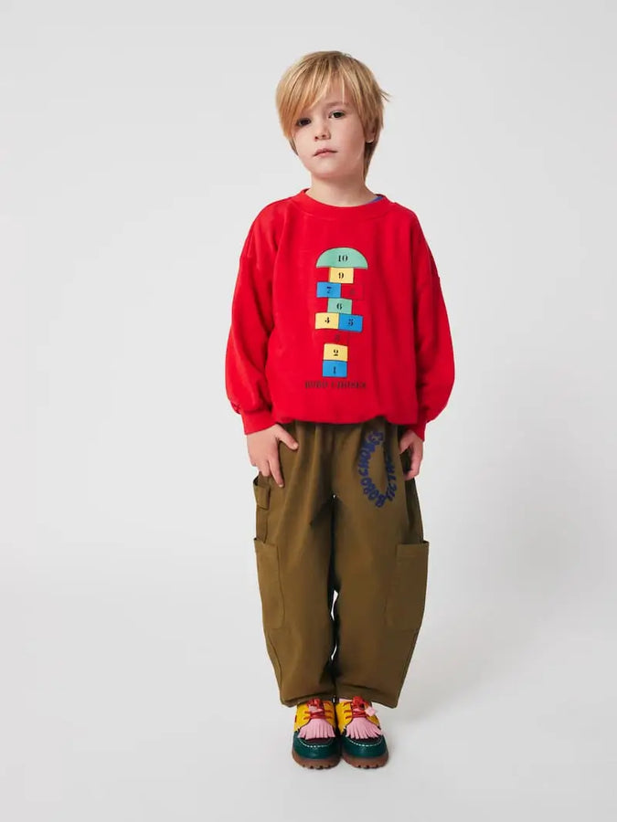 Niño vistiendo la sudadera roja con dibujo de rayuela y pantalones verde khaki de Bobo Choses.