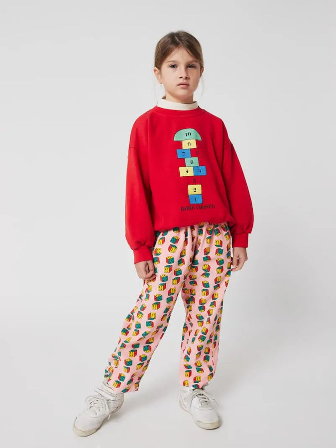 Niña vistiendo sudadera roja con dibujo de rayuela pantalones con estampado de dados multicolores.