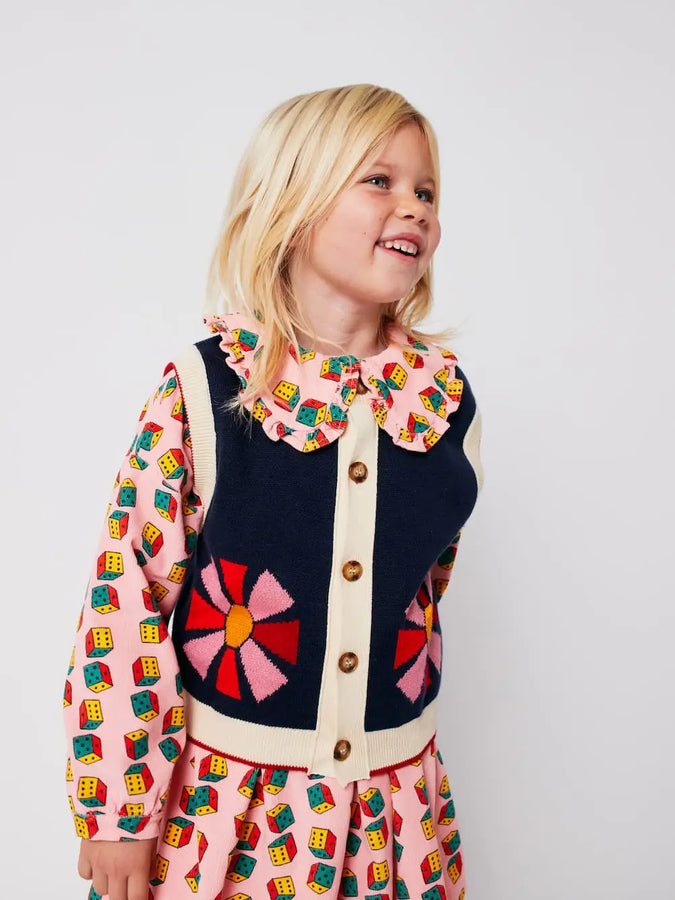 Niña con vestido rosa de dados multicolor, leotardos color block y chaleco de botones azul con dibujo de caleidoscopio, todo de Bobo Choses.