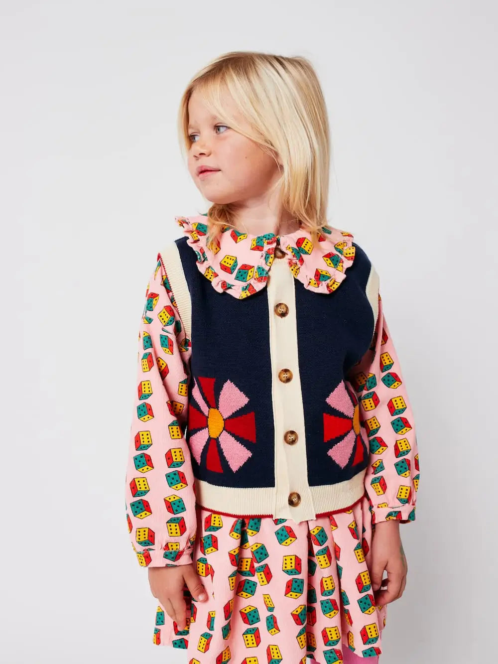 Niña con vestido rosa de dados multicolor, leotardos color block y chaleco de botones azul con dibujo de caleidoscopio, todo de Bobo Choses.