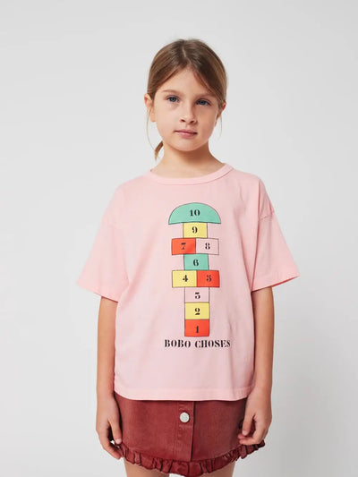 Niña vistiendo camiseta rosa con dibujo de rayuela y falda marron de Bobo Choses.