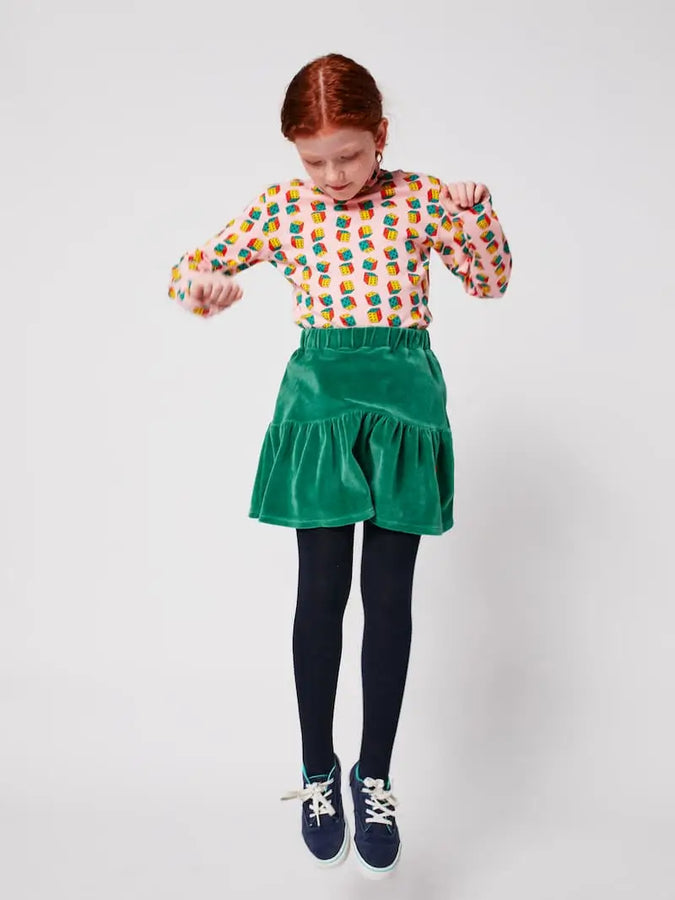Niña vistiendo falda verde de terciopelo y camiseta de cuello alto con mangas largas y estampado de dados de Bobo Choses.