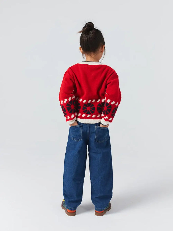 Niña posando de espaldas con pantalones vaqueros azules de Bobo Choses con corte cocoon y bolsillos traseros. Lo conjunta con jersey de color rojo y estampado de caleidoscopios oscuros también de Bobo Choses. 