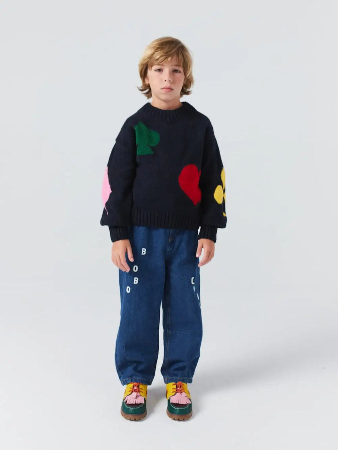 Niño con pantalones vaqueros azules con logo de Bobo Choses en la parte delantera con cintura ajustable y corte cocoon. Lo conjunta con jersey de punto de color azul marino y estampado de colores. 