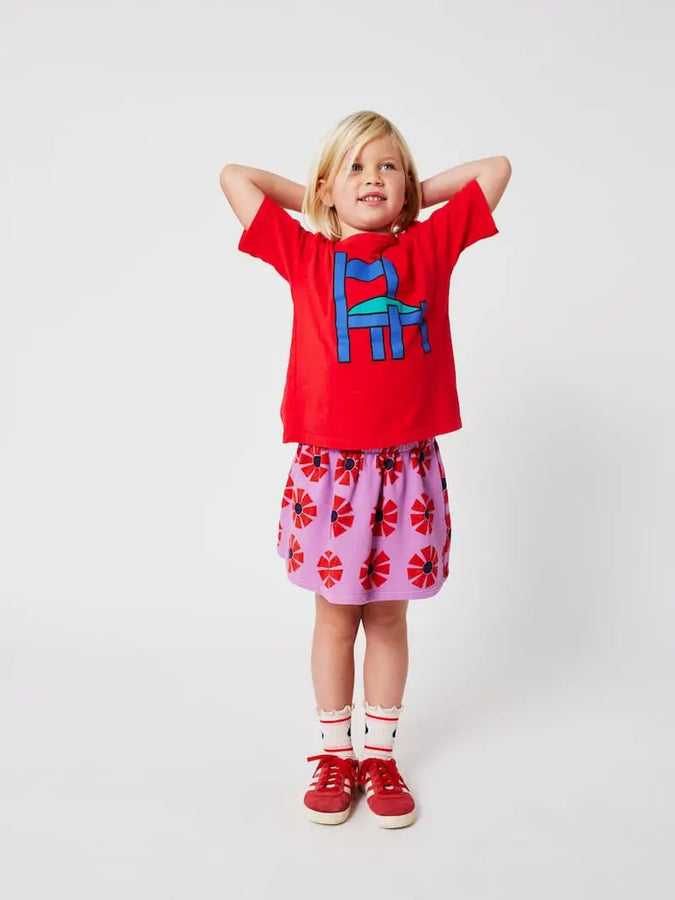Niña de frente con falda corta de Bobo Choses color morado con estampado de caleidoscopios rojos, con camiseta y zapatillas rojas a juego. 