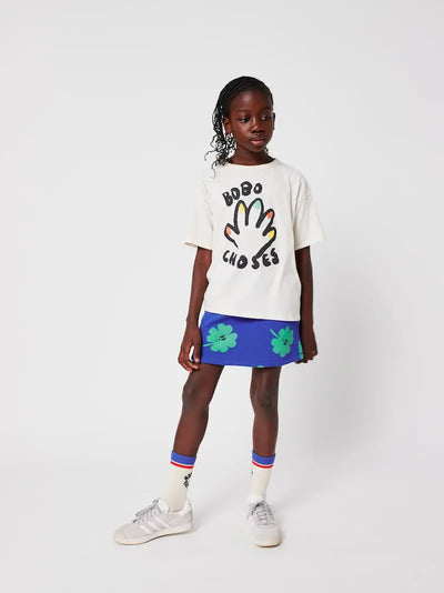Niña de color lleva una falda corta azul de Bobo Choses con estampado de tréboles verdes conjuntada con una camiseta de color blanco, calcetines blancos y deportivas grises. 