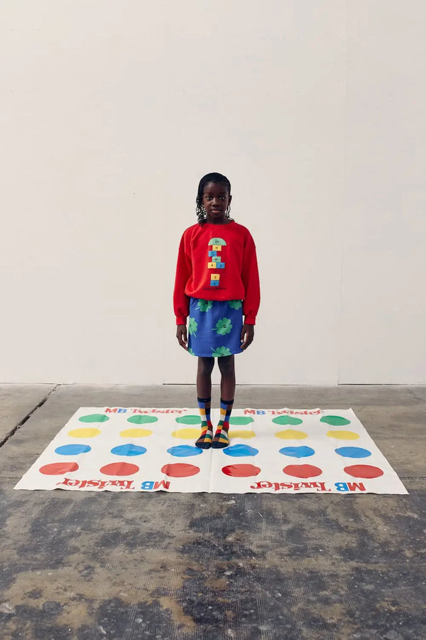 Niña de color lleva una falda corta azul de Bobo Choses con estampado de tréboles verdes conjuntada con una sudadera de color rojo. 