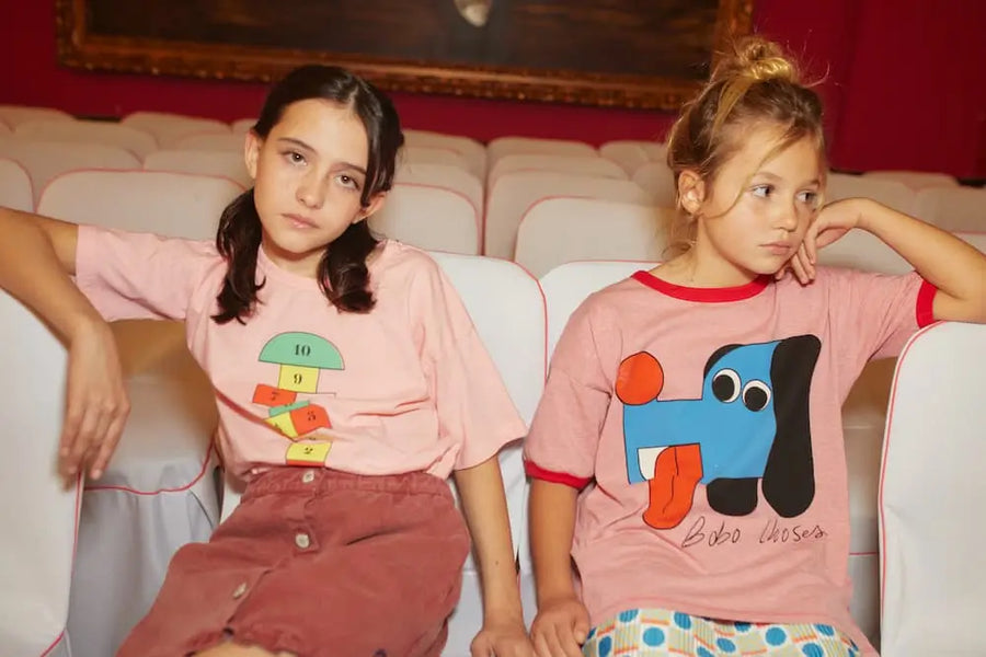 Dos niñas posando sentadas, una de ellas con una camiseta rosa de Bobo Choses Doggy Mate y otra con camiseta de dados de Bobo Choses.