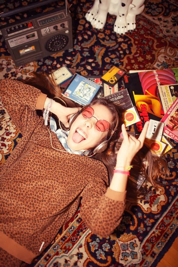 Niña sonriente tumbada en el suelo vistiendo sudadera Piupiuchick marrón de rizo con estampado de animal print y rodeada de cintas de casete 