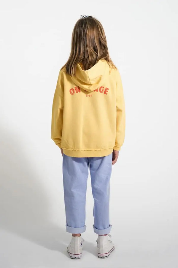 Vista atrás de niña con vaqueros azules y sudadera amarilla con capucha con texto On stage en la espalda.
